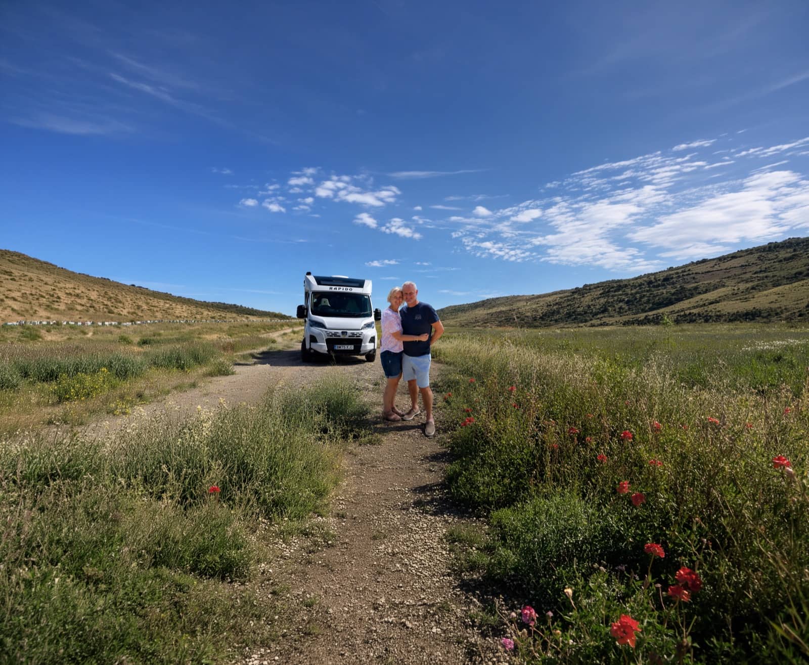 Et par som står ved siden av bobilen sin på en vei gjennom grønne, bølgende åser